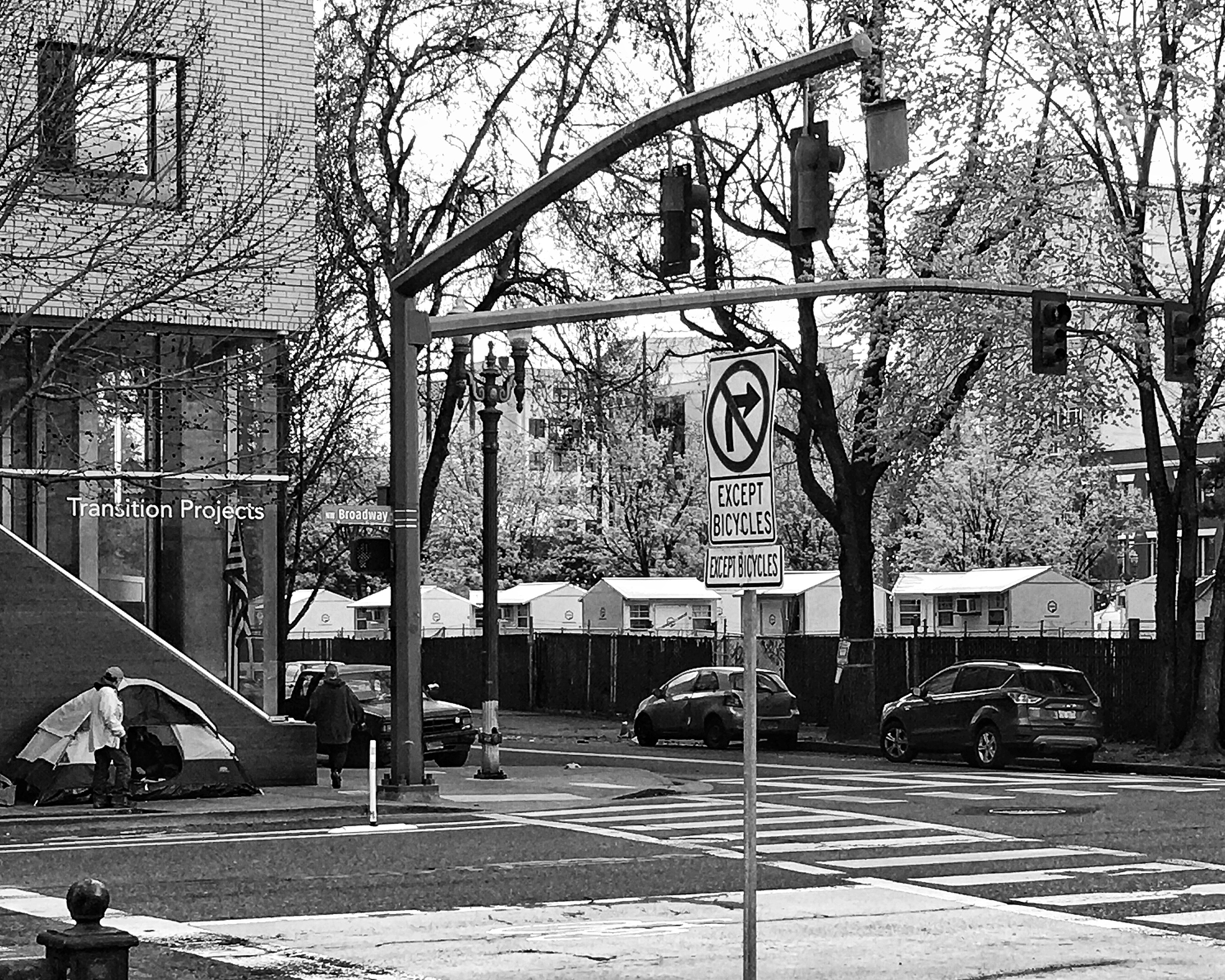 Black and white photograph of a makeshift food distribution box in front of a house in Portland Oregon containing cans of tomatoes, a bag of dried beans, and several onions.